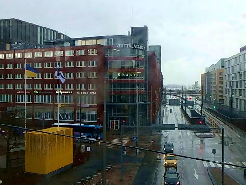 A rainy scene of red brick buildings and black asphalt. Some flag poles, one has the flag of Ukraine, the other has the State Flag of Finland.