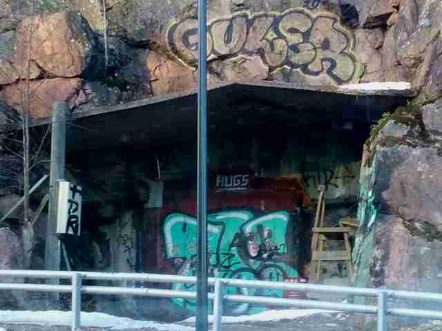 Kalliossa oleva metallinen ovi ja betoninen loiva katos. Paljon graffitia ympärillä, "guler" ja "hugs" selvimänä.